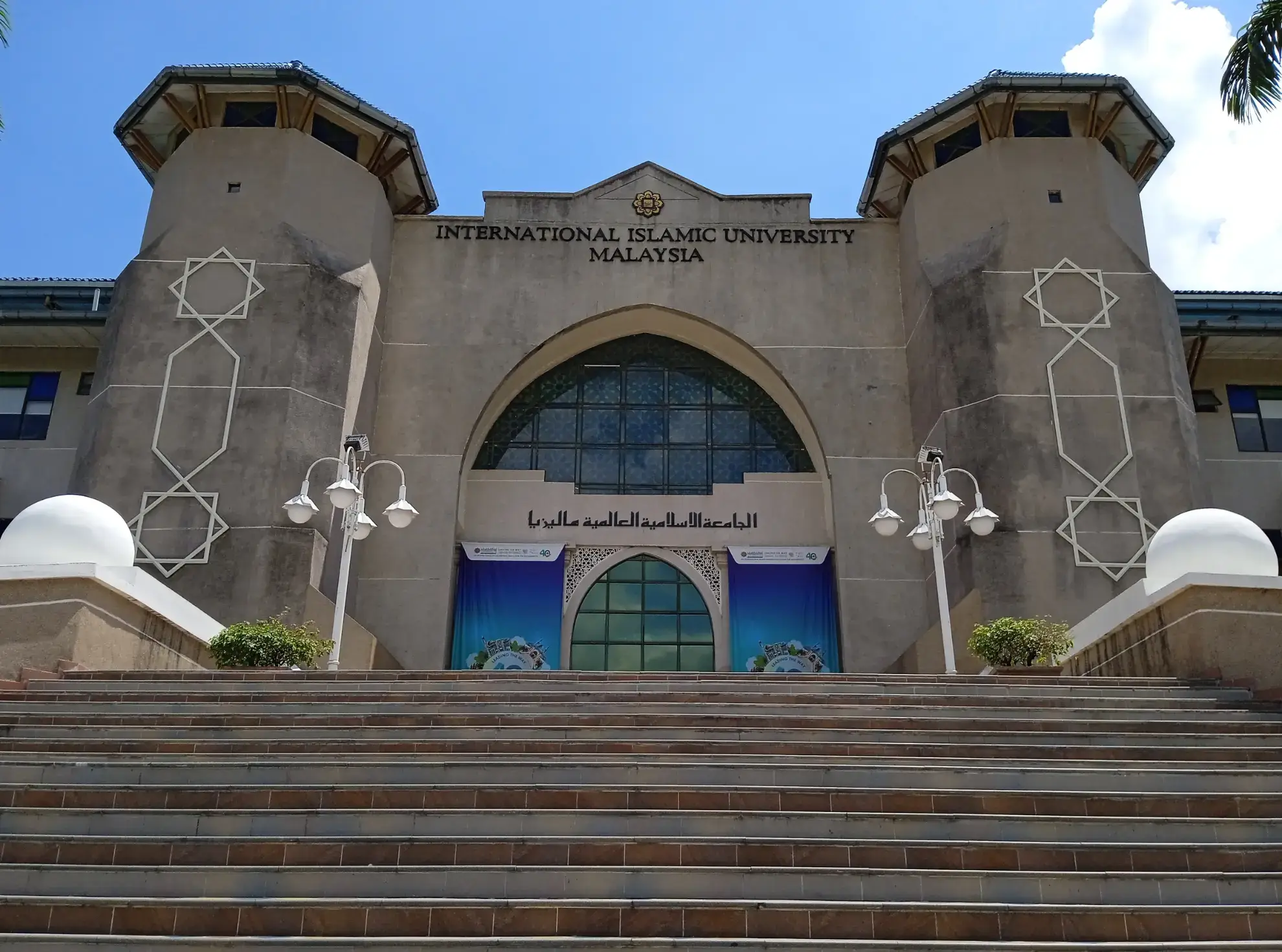 Stairs lead up to the entrance of International Islamic University Malaysia. The facade features tall towers and geometric patterns under a blue sky.