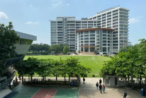 A large multi-story university building, typical of top universities in Malaysia, overlooks a green lawn and sports courts, with trees and people walking around the campus on a sunny day.