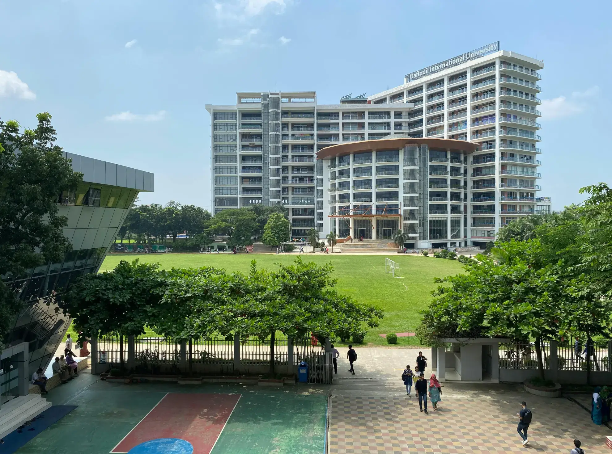 A large multi-story university building, typical of top universities in Malaysia, overlooks a green lawn and sports courts, with trees and people walking around the campus on a sunny day.