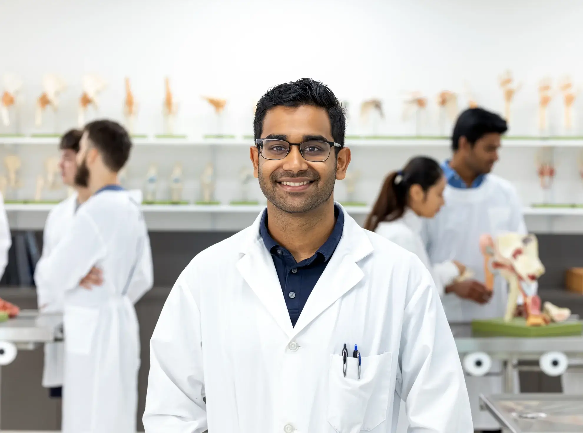 A person wearing a white lab coat and glasses stands smiling in a laboratory, with anatomical models and other people working in the background—an inspiring glimpse into the environment that awaits students pursuing an MBBS in Malaysia.